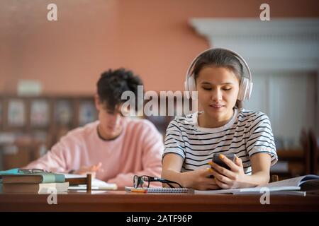 Grazioso studente universitario con cuffie a scorrimento in smartphone in biblioteca Foto Stock