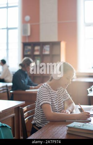 Ragazza adolescente diligente con cuffie ascoltare musica e prendere appunti Foto Stock