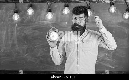 Uomo con barba e baffi sulla faccia di smemorati prurito testa. Barbuto hipster detiene orologio, lavagna su sfondo, copia dello spazio. Concetto di disciplina. Insegnante in occhiali tiene sveglia e penna. Foto Stock