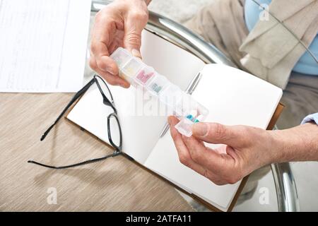 Mani di pensionato maschio che tiene il contenitore della pillola di plastica sopra il taccuino aperto Foto Stock