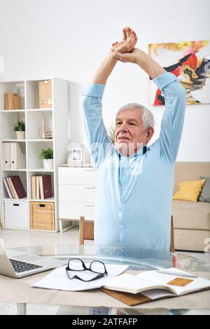 Uomo anziano leggermente stanco che alza le mani sopra la testa mentre si esercita da tavolo Foto Stock