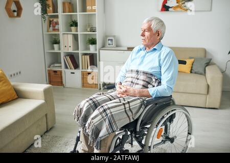 Triste e sereno senior disable uomo seduto in sedia a rotelle da divano a casa Foto Stock