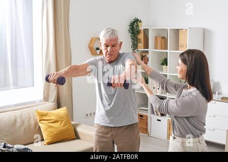 Giovane assistente sociale femminile che aiuta l'uomo anziano che esercita con i dumbbells Foto Stock