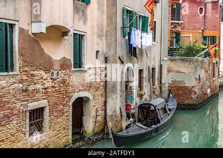 Gondola con tetto ormeggiato da una casa con persiane verdi e lavaggio appesa su una linea a secco e la bandiera veneziana volare fuori, Venezia, Italia Foto Stock