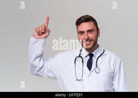 Bello moderno medico bianco stand medico in studio sfondo grigio pubblicità il tuo prodotto guardando la fotocamera e dito di puntamento via Positiv Foto Stock