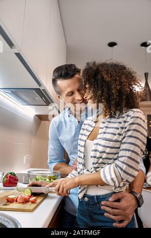 Data Romantica. Giovane coppia multietnica in piedi in cucina cucina cucina pranzo ragazzo abbracciando fidanzata tagliare verdure sorridente vista laterale felice Foto Stock