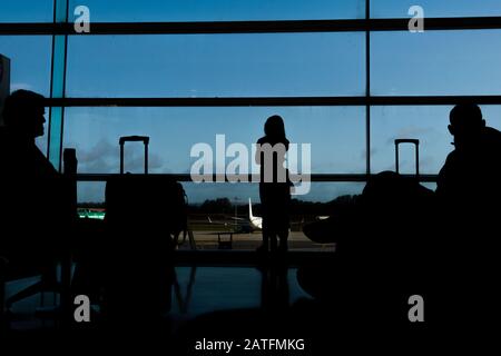 Sagoma dei passeggeri delle compagnie aeree in una sala d'aeroporto all'ampia finestra di osservazione che guarda un aereo che vola contro un tramonto surreale. Dublino Foto Stock