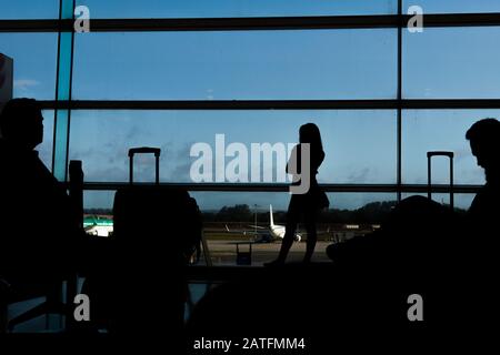 Sagoma dei passeggeri delle compagnie aeree in una sala d'aeroporto all'ampia finestra di osservazione che guarda un aereo che vola contro un tramonto surreale. Dublino Foto Stock