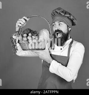 Concetto di dieta vegetariana. Uomo con barba su sfondo verde. Cuoco con il viso soddisfatto in uniforme borgogna tiene le verdure nel cestino di vimini. Lo chef tiene cavolo, ravanello, broccoli e carote. Foto Stock