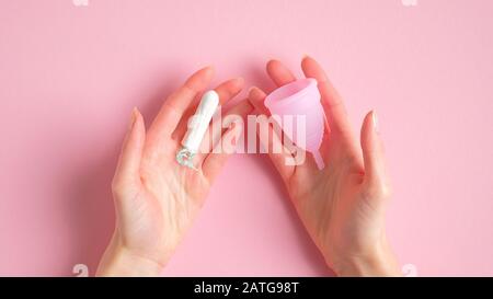 Mani femminili che tengono il tampone e la tazza mestruale sopra lo sfondo rosa. Confronto tra diversi tipi di prodotti per l'igiene mestruale. Giorni critici, mestruazioni Foto Stock