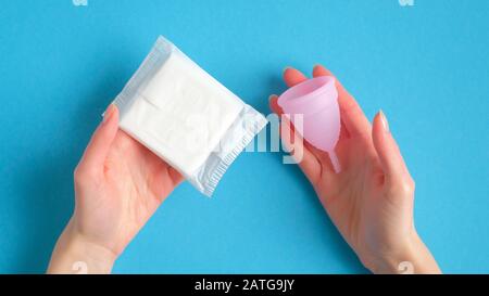 Le mani femminili che tengono la tazza mestruale e l'assorbente igienico sopra la priorità bassa blu. Diversi tipi di prodotti per l'igiene mestruale concetto di confronto Foto Stock