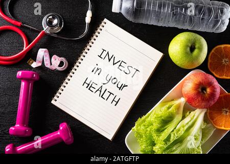 Stile di vita sano, concetto di cibo e sport. Vista dall'alto dello stetoscopio metro a nastro rosa manubrio, bottiglie d'acqua sportive, frutta e note book con Invest Foto Stock