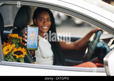 Giovane donna nera sorridente, che guida l'auto e mostra lo smartphone con l'applicazione di condivisione dell'auto Foto Stock