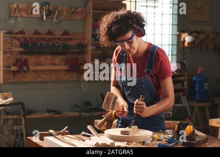 Donna curly concentrata in cuffie antirumore e occhiali di protezione utilizzando martello e scalpello mentre si lavora con asse di legno Foto Stock