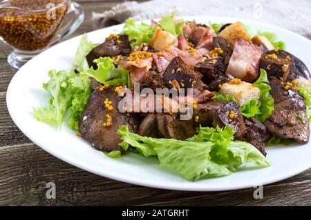 Insalata calda e sana di fegato di pollo, crostini di segale, pancetta affumicata, insalata verde e salsa di senape in un piatto bianco su sfondo di legno. Primo piano Foto Stock