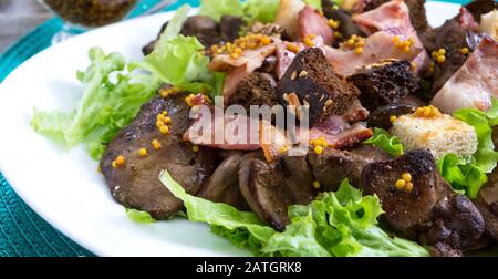 Insalata calda e sana di fegato di pollo, crostini di segale, pancetta affumicata, insalata verde e salsa di senape in un piatto bianco su sfondo di legno. Primo piano Foto Stock