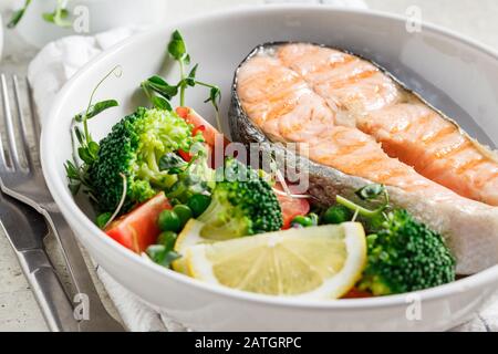 Bistecca di salmone alla griglia con broccoli e pomodori in un piatto bianco su sfondo grigio. Dieta alimentare concetto. Foto Stock