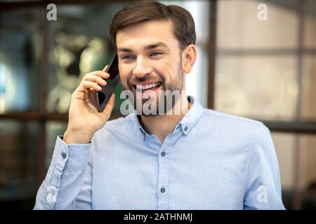 Giovane uomo bearded in una camicia blu che parla al telefono e che si sente impressionante Foto Stock