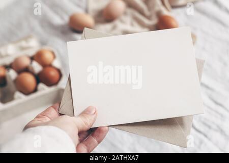 Primo piano della mano della donna che tiene carta bianca. Scenario di simulazione del biglietto di auguri. Primavera, design pasquale. Foto in borsa dallo stile femminile. Sfondo sfocato Foto Stock