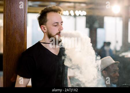 L'uomo fuma la narghilè nel bar. Primo piano di un ragazzo elegante per adulti che fuma una felpa con cappuccio al coperto di una caffetteria. Foto Stock