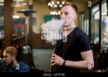 L'uomo fuma la narghilè nel bar. Primo piano di un ragazzo elegante per adulti che fuma una felpa con cappuccio al coperto di una caffetteria. Foto Stock