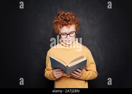 Ritratto di ragazzino in occhiali leggendo un libro contro lavagna Foto Stock