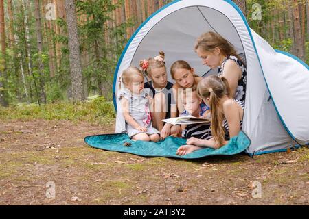 Madre che legge un libro seduto insieme in tenda con cinque bambini, mentre campeggio in una foresta estiva durante le vacanze estive Foto Stock