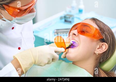 Ragazza bambino al medico. Il dentista posiziona un riempimento su un dente con la lampada di polimerizzazione dentale in cavità orale. Su sfondo clinico Foto Stock