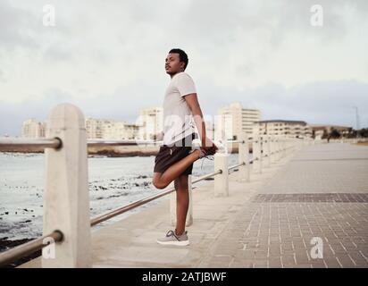 Vista laterale di un giovane atleta maschile runner che allunga le gambe in piedi accanto alla ringhiera sul lungomare Foto Stock