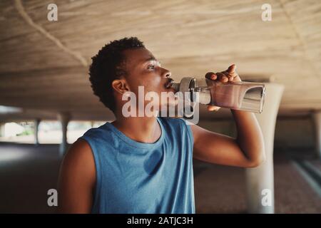 Vista laterale di un giovane uomo africano americano bere acqua da bottiglia in piedi sotto il ponte Foto Stock