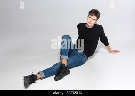 Elegante ragazzo che si posa in studio seduto sul pavimento in pantaloni denim e felpa nera. Ritratto maschile studente pieno su sfondo bianco. Studio Foto Stock