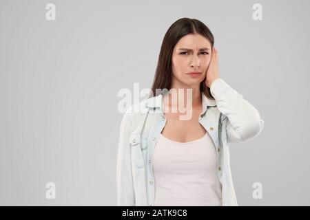 Giovane donna caucasica in maglia bianca su isolato grigio Sfondo Testa di toccare con mano con espressione dolorosa a causa del mal d'orecchi. Vista frontale di bruna guardando la fotocamera. Foto Stock