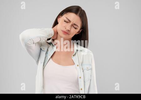 Giovane donna caucasica in maglia bianca su grigio Sfondo isolato collo di toccare con mano con espressione dolorosa a causa dell'Ache. Vista frontale della bruna con gli occhi chiusi. Foto Stock