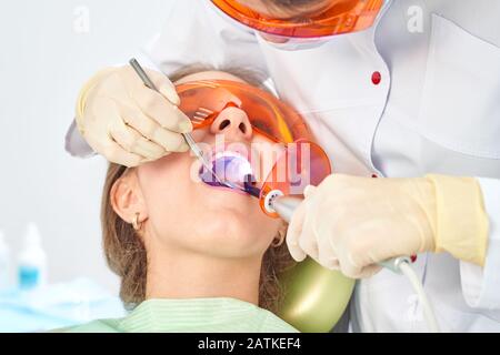 Ragazza bambino al medico. Il dentista posiziona un riempimento su un dente con la lampada di polimerizzazione dentale in cavità orale. Su sfondo clinico Foto Stock