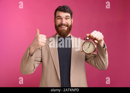 Felice giovane uomo d'affari che mostra il gesto dei pollici in su e tiene la sveglia sopra lo sfondo rosa Foto Stock