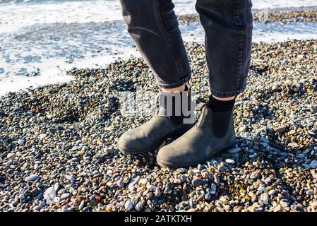 Stivali Chelsea classica suola in gomma nera. Concentrati sulle gambe di una donna hippster che indossa pantaloni neri denim. Sparato sulla spiaggia di ghiaia con il mare in grigro Foto Stock