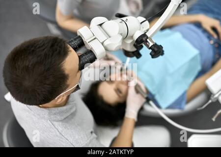 Il dentista in uniforme grigia con maschera protettiva e guanti in lattice bianco guarda al microscopio dentale mentre tratta il suo paziente femminile in una bib blu in A. Foto Stock