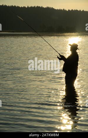 Pesca al mattino presto sul lago in estate. Silhouette del pescatore con canna da pesca in mano davanti alla luce del sole del mattino. Nel dista Foto Stock