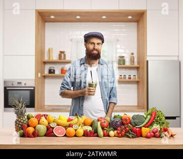 Giovane uomo informale che tiene un frullato verde in una cucina con frutta e verdure Foto Stock