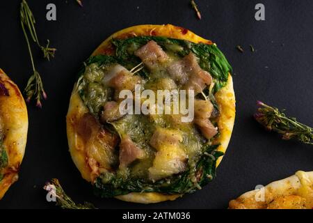 Piccola mini pizza. Pizza con pancetta, spinaci e formaggio su fondo nero. Pezzi di altre pizze sono visibili nelle vicinanze. Foto Stock