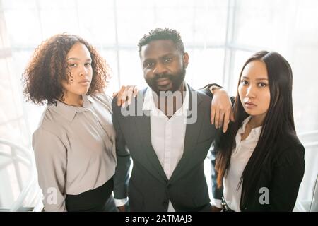 Ritratto di seri giovani colleghi multietnici in formalwear che si pongono insieme in ufficio Foto Stock