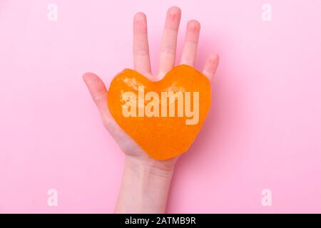 Arancio slime in forma di cuore nelle mani dei bambini. Ragazza mani che giocano il giocattolo di fango su sfondo rosa. Fare fango. Amore e concetto di giorno valentines Foto Stock