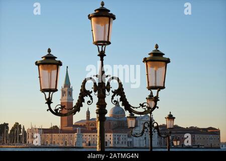 Due lampioni veneziani ornati, ciascuno con tre lanterne, incorniciano San Giorgio maggiore all'alba dal lungomare di San Marco a Venezia, in Italia Foto Stock