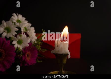 Candela corta che brucia sul pavimento in primo piano tra i fiori, accanto alla busta rossa contro sfondo scuro. Concetto romantico di tema serale Foto Stock