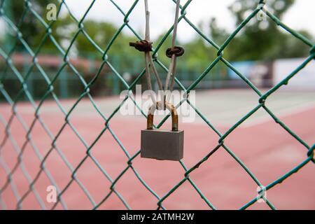 Primo piano colpo di arrugginito lucchetto su recinzione verde cablata. Campo da tennis in background, Foto Stock