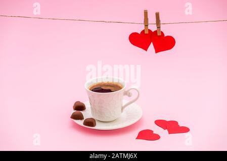 Tazza di chicchi di caffè con biscotti al cioccolato a forma di cuore e cuori di carta rossa su sfondo pastello rosa. Appendiabiti e cuori di carta rossa sulla corda. Va Foto Stock