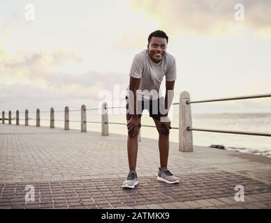 Ritratto di un uomo giovane e sorridente stanco che prende pausa dopo aver fatto jogging sul lungomare Foto Stock