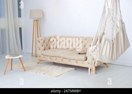 interno soggiorno con amaca, soffice tappeto e tavolino da caffè con lampada. Appartamento loft multifunzionale con amaca, divano beige e grande finestra Foto Stock