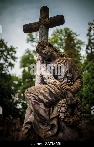 Vecchia scultura in pietra di donna contemplativa appoggiata alla croce nello storico cimitero di Rakowicki a Cracovia, Polonia Foto Stock