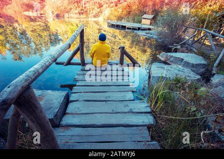 Un uomo seduto su un ponte di legno e guardando un lago di foresta con una riva di granito. Alba sul lago in autunno Foto Stock